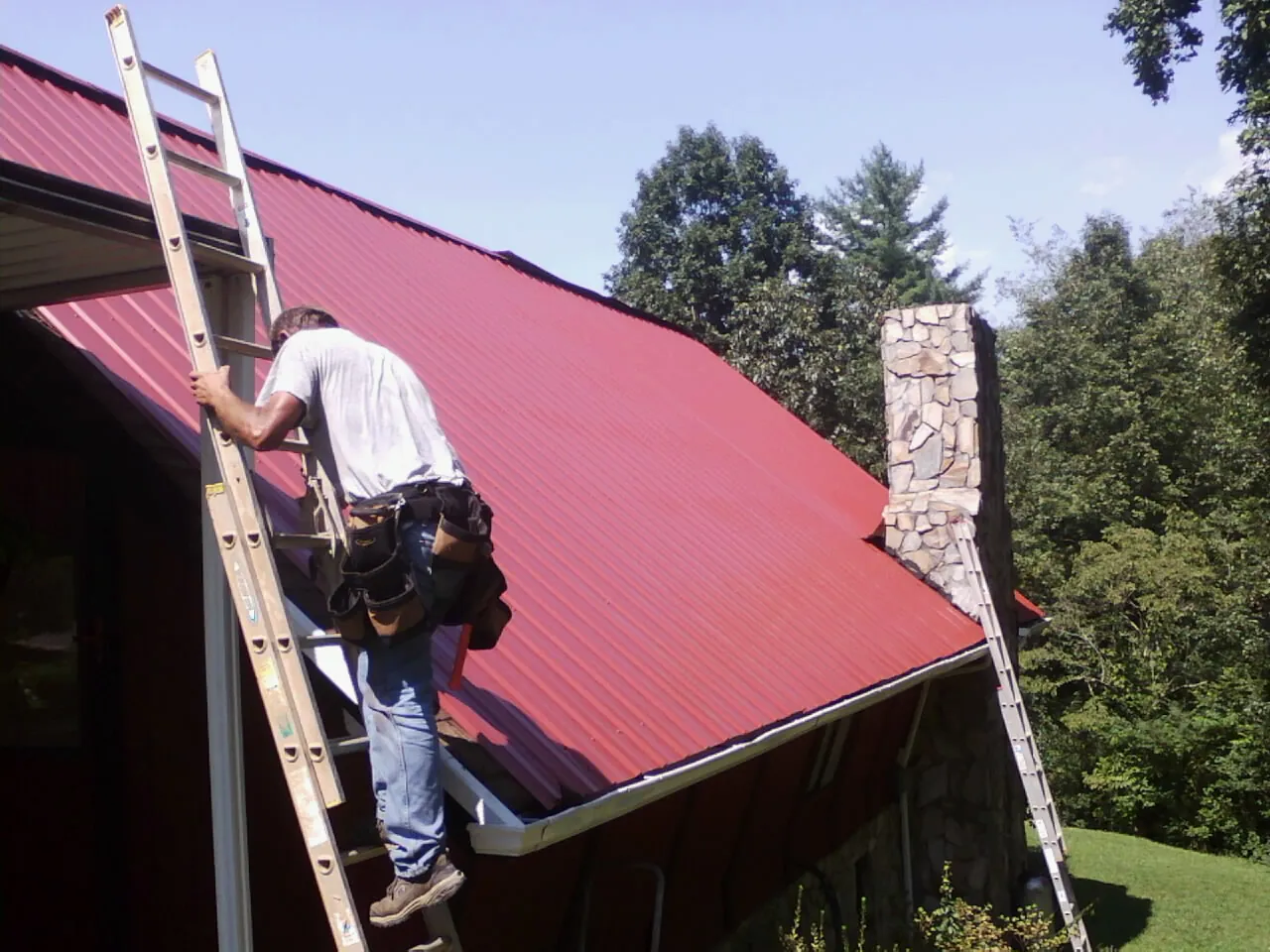 Roofer on a ladder beside a red metal roof and stone chimney — Soffit & Fascia Repair by Roofing Brothers in Amawalk